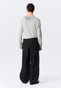 Homme de dos portant une chemise à manches longues gris clair et un pantalon cargo noir ample avec de grandes poches, sur un fond blanc.