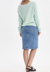 Pull en tricot vert menthe, jupe crayon en denim bleu avec ourlet effiloché, et sandales plates noires. Vue de dos mettant en valeur les détails de la tenue.