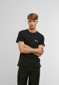 Sort bomulds T-shirt med rund halsudskæring, korte ærmer og et lille broderet hvidt logo på venstre brystområde.