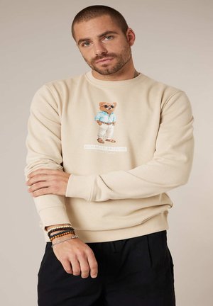 Man met kort haar en baard, draagt een beige sweatshirt met een berenafbeelding en tekst, en heeft kralen- en kettingarmbanden om zijn linkerpols.