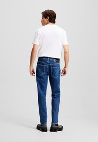 Jean en denim bleu avec une coupe droite, associé à un t-shirt blanc uni. Les caractéristiques notables incluent une étiquette noire sur la taille et deux poches arrière.
