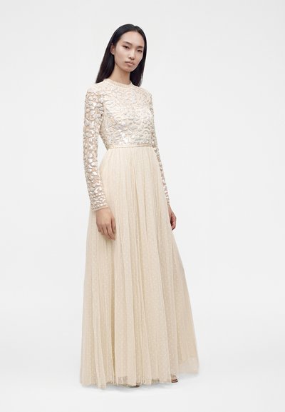 Femme portant une longue robe beige avec un motif de sequins sur le haut et une jupe transparente à pois, debout face à l'avant avec une expression neutre.