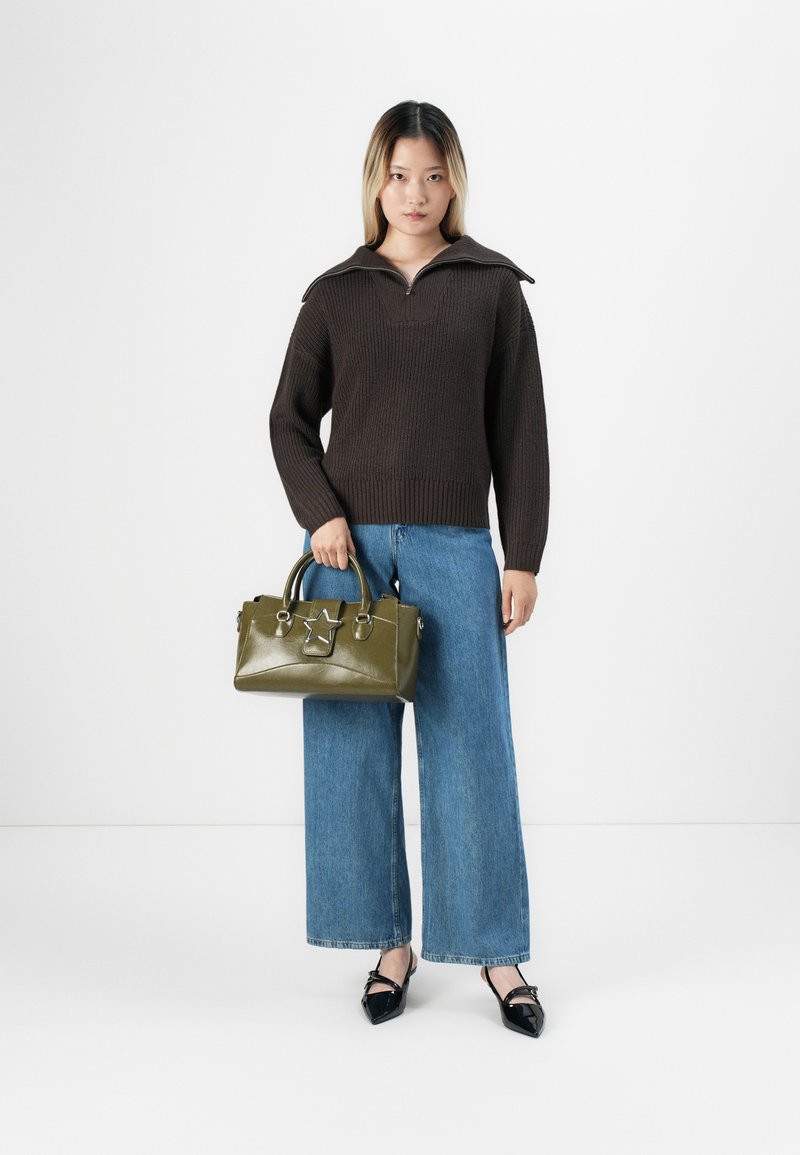 Femme portant un pull en maille brun foncé et un jean bleu à jambes larges, tenant un sac à main olive avec un détail en forme d'étoile, debout sur fond blanc.