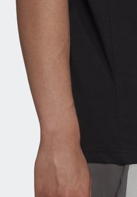 En vuxens underarm och hand bredvid en svart kortärmad skjorta och grå byxor, visad mot en enkel ljus bakgrund.