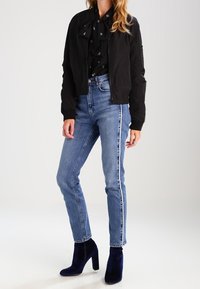 Veste bomber noire portée sur un chemisier noir avec un nœud, associée à un jean taille haute bleu clair avec des bandes latérales et des bottines bleu marine.