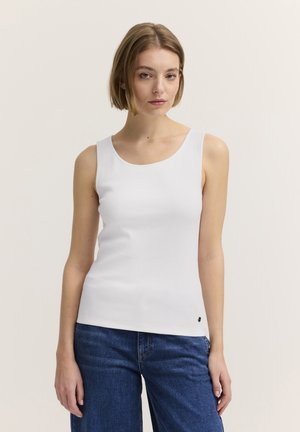 Junge Frau mit kurzen Haaren, die ein weißes ärmelloses Top und blaue Jeans trägt, steht vor einem schlichten hellen Hintergrund.