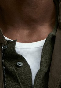 Olivengroene, gebreide jas met ritssluiting en gestructureerde stof, gedragen over een wit geribd T-shirt. Voorzien van ronde zwarte knopen en een hoge kraag.