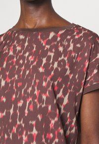 Primer plano de una persona que lleva una blusa de patrón abstracto en marrón y rojo, con mangas cortas y un escote redondo.