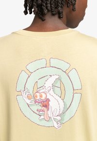 Geel katoenen t-shirt met een grote, kleurrijke graphic van een karakter met overdreven kenmerken, tegen een circulair ontwerp.