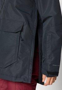 Temno modra vodoodporna jakna z zadrgo na sprednji strani, večjim številom žepov, nastavljivimi manšetami in gladko, mat površino ter detajlom logotipa.