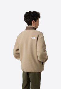 Noor inimesel on lokkis juuksed ja ta kannab beeži fleece-tüüpi jopet pruunide krae ja The North Face'i logoga, käed taskutes, seljaga vaataja poole.