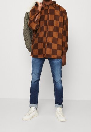 Chemise à carreaux marron et beige avec une coupe décontractée, superposée sur une veste verte. Jean bleu avec les poignets retroussés et baskets blanches.