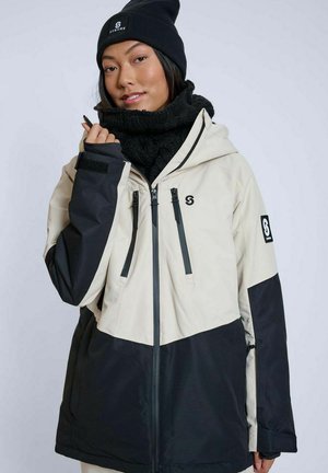 Kvinde iført sort hue og tørklæde, beige og sort jakke med lynlåse og logoer, poserer med hånden tæt på ansigtet mod ensfarvet baggrund.