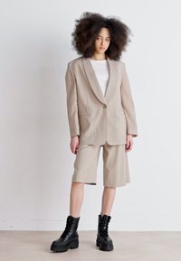 Blazer oversize beige strutturato con un solo bottone, abbinato a pantaloni beige. Il modello indossa stivali neri con lacci spessi. Lo sfondo è neutro.
