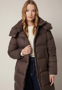 Brązowa kurtka puchowa z wysokim kołnierzem, zatrzaskami i bocznymi kieszeniami. Gładka tekstura i pikowana konstrukcja zapewniają ciepło i komfort.