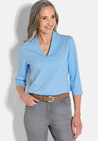 Hellblaues V-Ausschnitt-Shirt aus glattem Stoff, mit Dreiviertelärmeln, kombiniert mit grauen Jeans und einem braunen Gürtel.