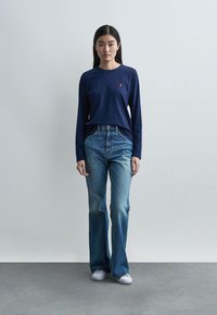 Γυναίκα στέκεται μπροστά σε απλό φόντο φορώντας μπλε πουκάμισο με μακριά μανίκια, ψηλόμεσα μπλε τζιν και λευκά αθλητικά παπούτσια, κοιτάζοντας προς τα εμπρός.