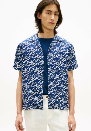 Joven con una camisa azul de manga corta estampada sobre una camiseta azul marino, pantalones blancos, posando sobre un fondo liso.