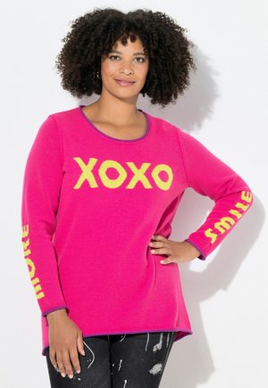 Žena s kudrnatými vlasy, oblečená ve světlém růžovém svetru se žlutým nápisem "XOXO" na hrudi a "smile" na rukávech, pózující s jednou rukou na bocích.