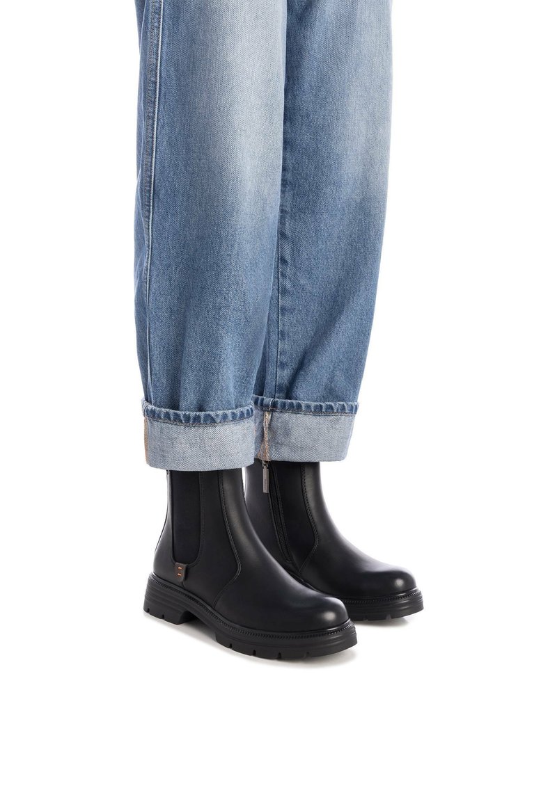 Stivaletti neri in pelle, dotati di pannelli elastici laterali, una suola in gomma testurizzata e una punta arrotondata. Indossati con jeans di denim blu risvoltati.