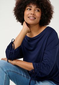 Haut bleu marine à encolure large et manches trois-quarts, avec des détails noués aux poignets. Tissu doux et lisse avec une coupe décontractée.