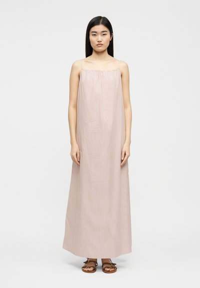 Γυναίκα με ίσια μαύρα μαλλιά, που φοράει ένα αμάνικο φόρεμα ριγέ σε ανοιχτό ροζ και λευκό μέχρι τον αστράγαλο, και καφέ σανδάλια, στέκεται μπροστά σε ένα λευκό φόντο.