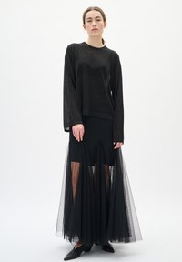 Haut à manches longues noir texturé associé à une jupe noire avec des panneaux en tulle transparent, créant un effet superposé. Fini lisse, ajusté au corps.