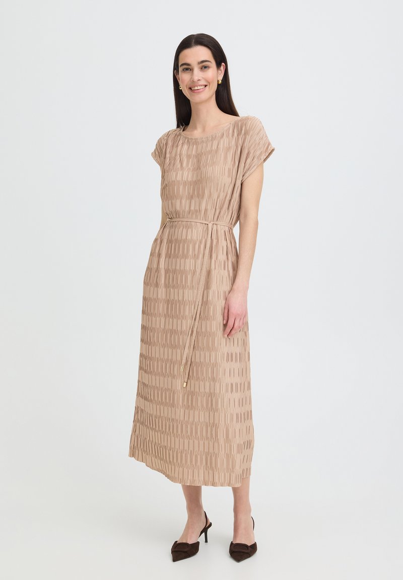 Femme aux longs cheveux bruns souriante, portant une robe midi beige texturée avec une ceinture à la taille et des escarpins pointus foncés, debout devant un fond uni.