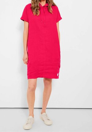 Femme portant une robe à capuche à manches courtes rose vif avec cordons, associée à des baskets beige clair, debout devant un fond uni.