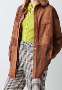 Brązowa zamszowa koszula typu overshirt z dwoma kieszeniami na piersi, nałożona na jasną zieloną prążkowaną bluzkę i czarno-białe kraciaste spodnie. Widoczne detale z guzikami.