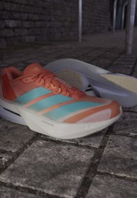 Jaskrawo pomarańczowe i jasno niebieskie buty do biegania z białą podeszwą spoczywające na brukowanej nawierzchni przy słabym oświetleniu.