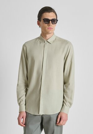 Joven con gafas de sol oscuras, camisa beige clara de manga larga abotonada y pantalones grises, de pie frente a un fondo liso.