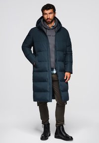 Pufferjas, donkerblauwgroen, knielengte, gequilt ontwerp met een capuchon; gedragen over een grijze hoodie, gecombineerd met olijfgroene broek en zwarte veterschoenen.
