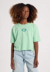 Lichtgroene cropped t-shirt gemaakt van katoen, met een circulair logo met de tekst "California Malibu" in blauw en geel. Zachte textuur, korte mouwen.