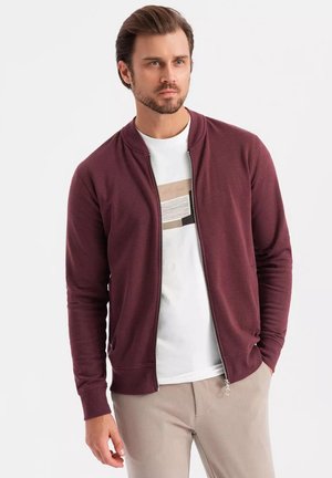 Chaqueta burdeos con cremallera hecha de una tela suave, con puños acanalados, llevada sobre una camiseta gráfica blanca, combinada con pantalones claros.