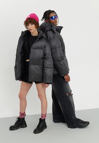 Svarte pufferjakker med en blank finish, kombinert med svarte shorts. Den ene bærer en rosa lue og rosa stripete sokker; den andre har solbriller.