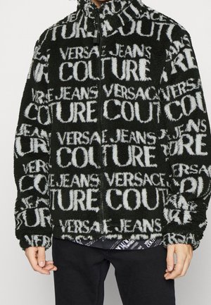 Veste zippée noire et blanche duveteuse avec le texte répété "Versace Jeans Couture", portée par une personne avec un pantalon noir.