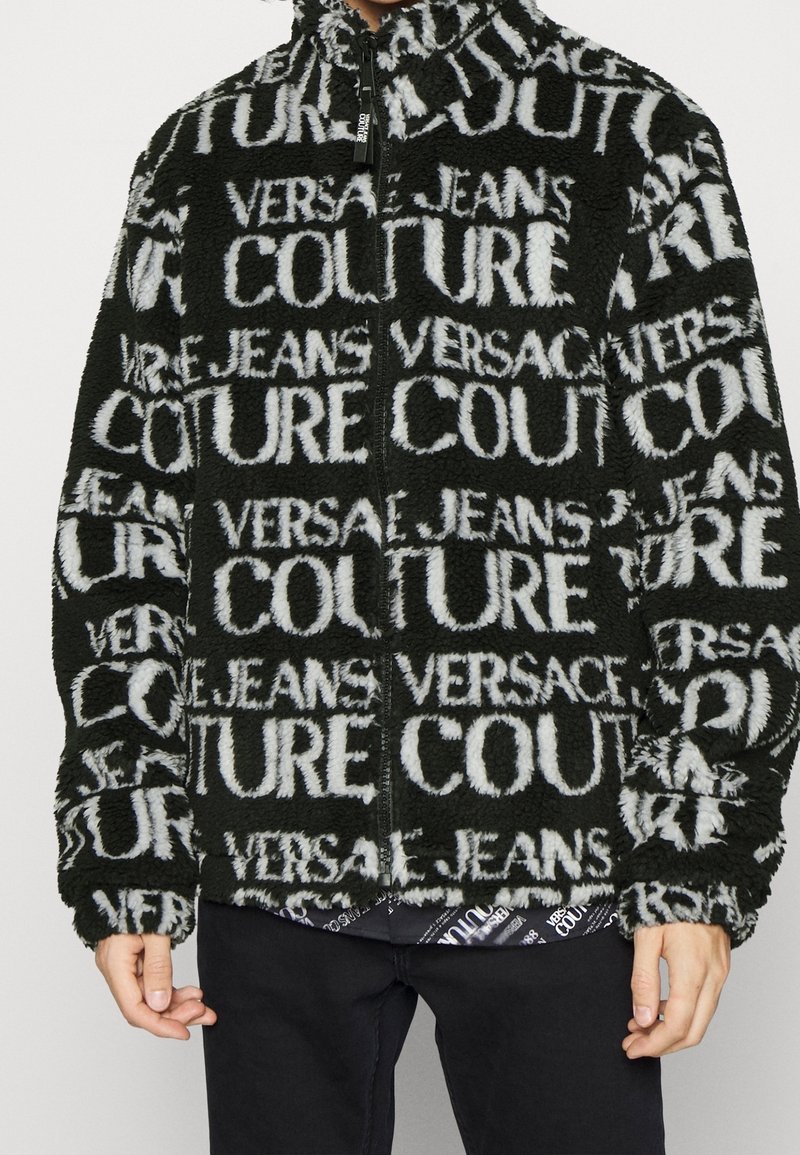 Veste zippée noire et blanche duveteuse avec le texte répété "Versace Jeans Couture", portée par une personne avec un pantalon noir.