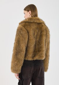 Brun faux fur jakke med krave, cropped design og lange ærmer, parret med mørke bukser, som fremviser en blød, tekstureret finish.
