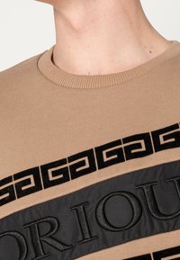 Felpa beige con collettino a coste, fascia in velluto nero con testo stampato in rilievo e striscia di tessuto nero a contrasto con motivo geometrico.