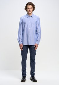 Camisa de botones azul claro con un bolsillo, de mangas largas y un corte relajado, combinada con pantalones azul oscuro y zapatos negros.