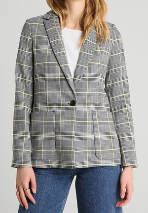 Žena oblečená v šedém kostkovaném blejzru s jedním černým knoflíkem přes bílé tričko a modré džíny, stojí s uvolněnýma rukama podél těla.