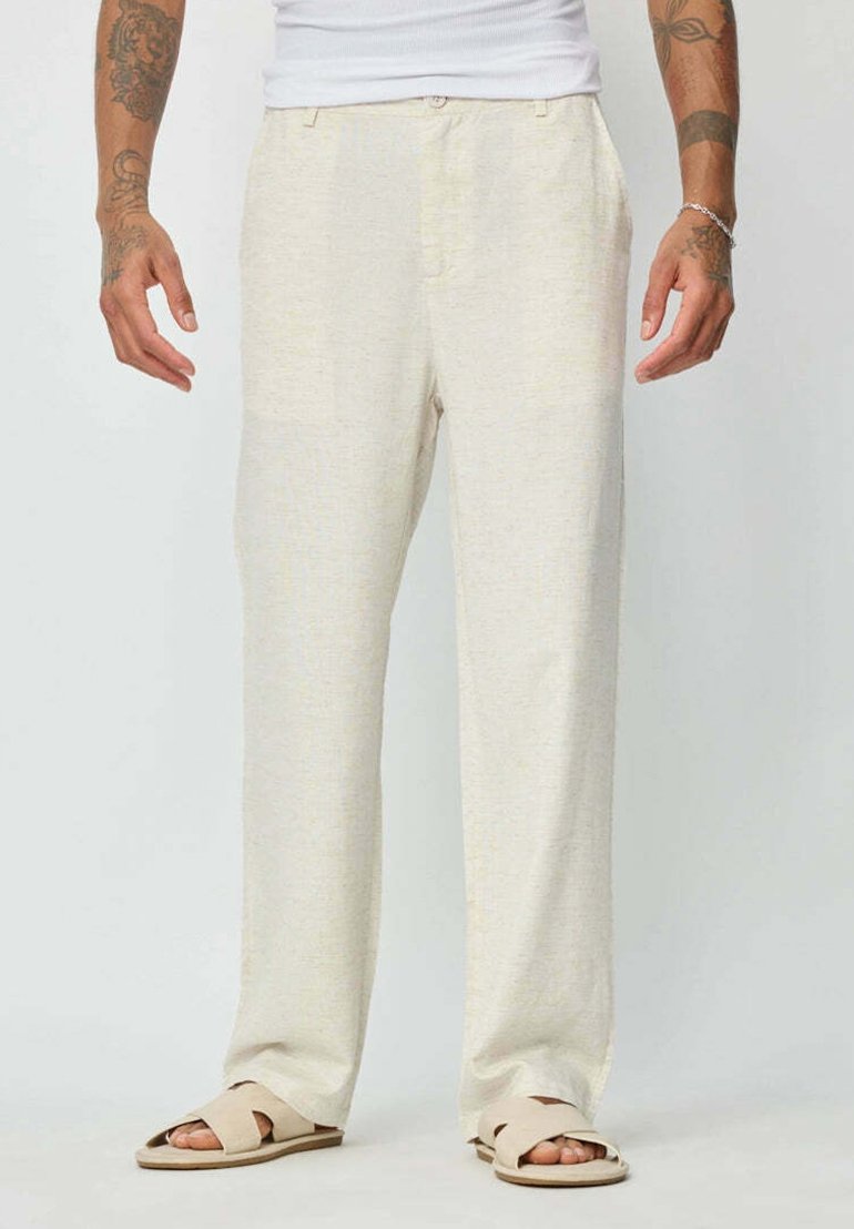 Pantalons en lin beige clair avec une coupe décontractée, présentant un devant plat, des poches latérales et une texture subtile. Portés avec des sandales crème.