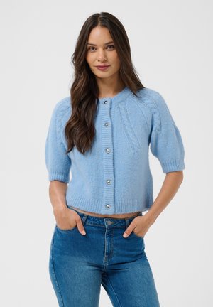 Giovane donna con capelli lunghi e castani che indossa un cardigan azzurro chiaro a maniche corte e jeans blu, in piedi con le mani in tasca.