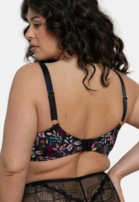 Femme aux cheveux bouclés portant un soutien-gorge floral avec des bretelles noires ajustables et des sous-vêtements en dentelle noire, se tenant avec une main sur la hanche, dos visible.