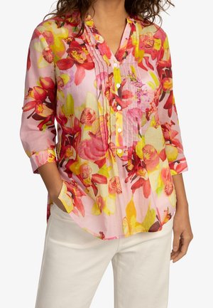 Femme portant un chemisier rose à fleurs à manches longues avec boutons et plis, associé à un pantalon blanc, mains dans les poches.