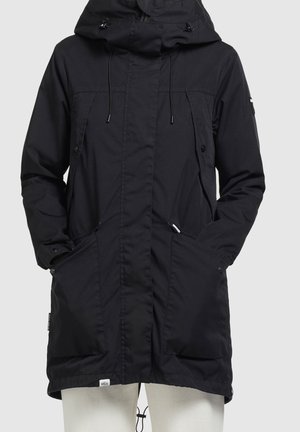 Czarna długa kurtka z kapturem, z przednią klapą, troczkami i dużymi ukośnymi kieszeniami, noszona z rękami włożonymi do kieszeni.