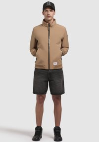 Chaqueta cremosa con cierre y cuello alto, con acentos negros y bolsillos laterales, combinada con pantalones cortos oscuros y zapatillas negras.