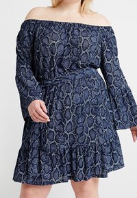 Kvinna som bär en off-shoulder långärmad blå klänning med ormskinnsmönster, midjefäste och volangnedkant, stående mot en enfärgad bakgrund.