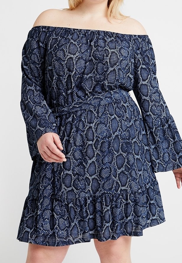 Kvinna som bär en off-shoulder långärmad blå klänning med ormskinnsmönster, midjefäste och volangnedkant, stående mot en enfärgad bakgrund.